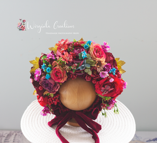 Colorful handmade floral bonnet for newborn photography, featuring rich red, pink, burgundy, blue, and yellow artificial flowers on a wooden mannequin head. Tied with a velvet ribbon, perfect for photo shoots and baby portraits. Wirginta Creations the creator.