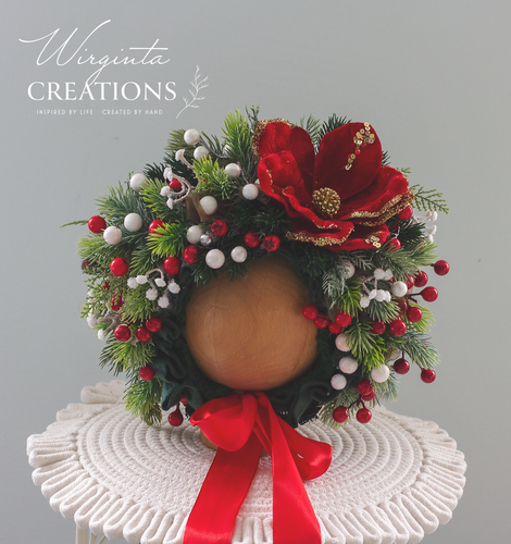 Christmas-themed baby headpiece with artificial red flower, berries, pinecones, and greenery, arranged around a rounded base with red ribbon bow. Designed for short-term wear and seasonal photography for babies aged 6–24 months.