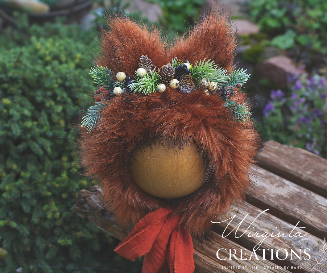 Fox bonnet, burnt orange colour, decorated with faux fur and artificial greenery. Placed on a wooden ball stand, green-outdoor background. Photography Prop. Creator-Wirginta Creations