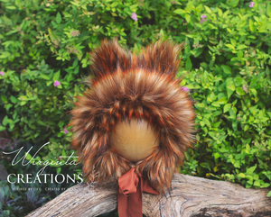 Fox inspired bonnet decorated with faux fur placed on a wooden ball standing on a log. Burnt orange colours, fabric tie. Green bush background. Bonnet created by Wirginta Creations