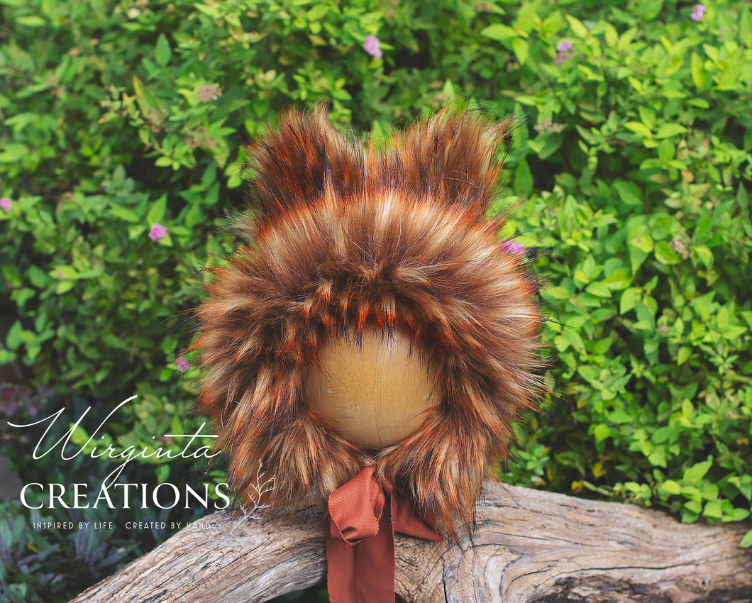 Fox inspired bonnet decorated with faux fur placed on a wooden ball standing on a log. Burnt orange colours, fabric tie. Green bush background. Bonnet created by Wirginta Creations