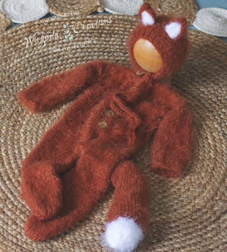 Knitted burnt orange baby outfit with white accents on a textured surface. Knitted fox-inspired footed romper with tail and matching fox bonnet presented on wooden ball.