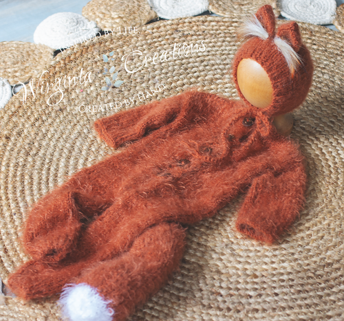 Knitted burnt orange baby outfit with white accents on a textured surface. Knitted fox-inspired footless romper with tail and matching fox bonnet presented on wooden ball.